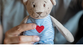 Peluche en forme d’ours tenue dans une main, portant un vêtement rayé bleu et blanc avec un cœur rouge, symbolisant la tendresse et l’enfance.